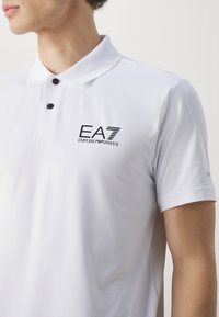 Weißes Polohemd aus atmungsaktivem Stoff mit strukturiertem Muster. Mit drei schwarzen Knöpfen und dem Logo 'EA7 EMPORIO ARMANI' auf der Brust.