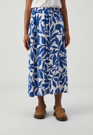 Person iført en hvid overdel, blå og hvid blomsterprintet nederdel og brune lukkede sandaler står foran en ensfarvet baggrund.