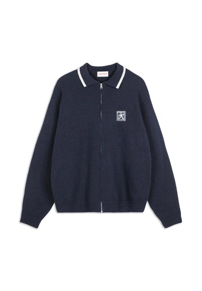 Marinblå stickad zip-up tröja med vit rand på kragen och liten broderad patch på vänster bröst.