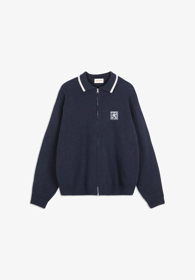 Marinblå stickad zip-up tröja med vit rand på kragen och liten broderad patch på vänster bröst.