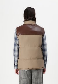 Beige Puffer-Weste mit einem braunen Kunstleder-Einsatz. Sie verfügt über ein gepolstertes Design, einen hohen Kragen und wird über einem blauen karierten Hemd getragen.