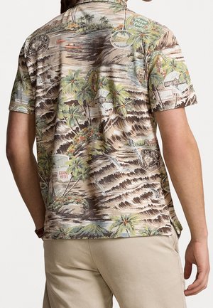 Man draagt een shirt met korte mouwen met tropische print van palmbomen, bergen, hotelborden en meeuwen, gecombineerd met lichtbeige broek.
