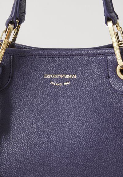 Navy blue leather handbag features a gold Emporio Armani logo with "MILANO 1981" underneath, highlighting craftsmanship and luxury.