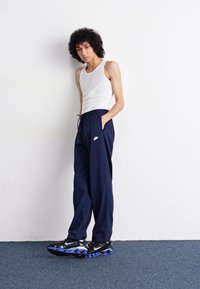 Mørkeblå joggebukser med elastisk liv, Nike-logo, sammen med en hvit tanktop og svarte joggesko med blå detaljer.