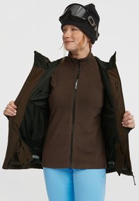 Bruine fleece jack, geopend, met een zwarte binnenvoering zichtbaar. Het model draagt een zwarte beanie en skibril, met blauwe broeken zichtbaar daaronder.