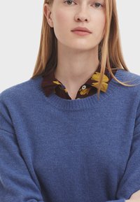 Ljusblå tröja i texturerad stickad kvalitet, med rund halsringning och sänkta axlar, lagd över en blommönstrad skjorta.