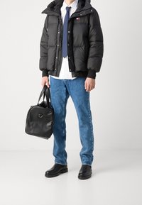 Schwarze Pufferjacke, blaue Jeans, schwarze Schuhe, weißes Hemd, marineblauer Krawatte und eine schwarze Lederhandtasche mit runder Form und oberen Griffen.