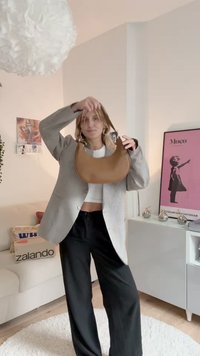 Brown leather hobo bag with a curved shape and silver hardware, held by a person wearing a grey checked blazer and black pants.