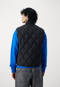 Schwarze gesteppte Weste mit einem Muster aus erhabenen, ovalen Formen, getragen über einem blauen Langarmshirt und grauen Jeans, von hinten betrachtet.