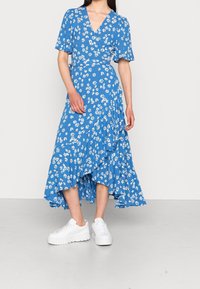 Robe portefeuille bleue à fleurs avec des manches courtes, ourlet froncé et baskets blanches. Le tissu présente de petites fleurs blanches sur un fond texturé.