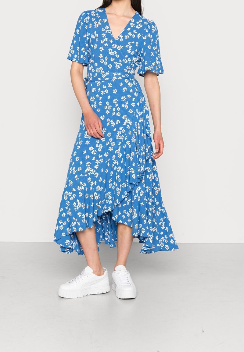 Robe portefeuille bleue à fleurs avec des manches courtes, ourlet froncé et baskets blanches. Le tissu présente de petites fleurs blanches sur un fond texturé.