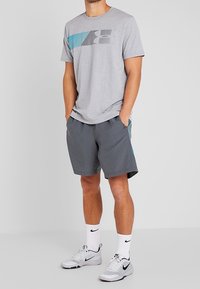 Muž oblečený v šedém grafickém tričku, tmavě šedých šortkách s bočním detailem, bílých ponožkách Nike a bílých teniskách Nike, stojící s rukama v kapsách.