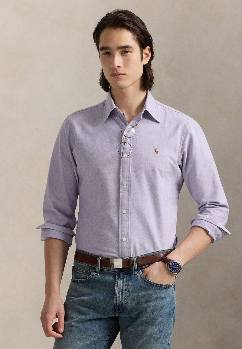 Ανοιχτό μωβ πουκάμισο με μακριά μα νίκια και κουμπιά, με μικρό κεντημένο λογότυπο, συνδυασμένο με τζιν και καφέ ζώνη. Περιλαμβάνει στρογγυλά γυαλιά και ρολόι.