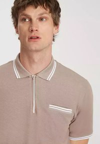 Beżowa koszulka polo z krótkim rękawem, z zamkiem przy kołnierzyku i białymi paskami. Posiada małą kieszonkę z białym akcentem. Miękka tekstura.