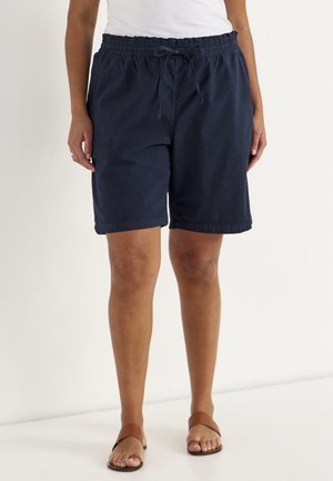 Person iført marineblå elastiske shorts med snøre i taljen, hvid top og brune sandaler, stående mod en hvid baggrund.