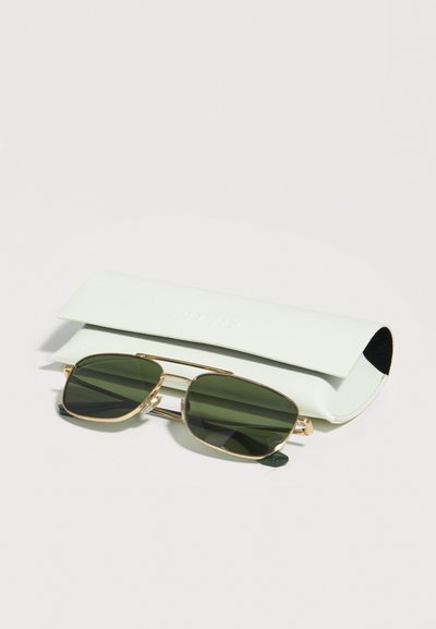 Ορθογώνια γυαλιά ηλίου με πράσινους φακούς και λεπτό χρυσό σκελετό, τοποθετημένα μπροστά από μια κλειστή λευκή δερμάτινη θήκη γυαλιών πάνω σε λευκή επιφάνεια.