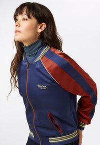 Navy bomber jacket with red satin sleeves, striped ribbed cuffs, and gold embroidery. Features zip closure and side pockets.