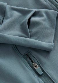 Fleecejakke i en dæmpet blå-grå farve, med en glat tekstur, fold-over krave og en lynlåslomme med kontrastfarvet trækhank.