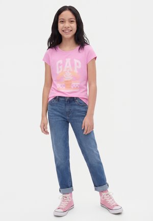 Mädchen trägt ein pinkes GAP-T-Shirt mit Eiscreme-Motiv, hochgekrempelte blaue Jeans und rosa High-Top-Sneaker, steht und lächelt.