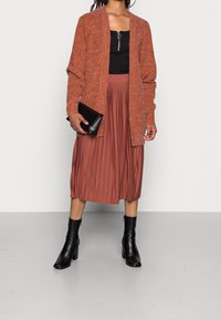 Cardigan lavorato a maglia color ruggine, top aderente nero, gonna midi plissettata in arancio bruciato, pochette nera e stivaletti neri con tacco alto.