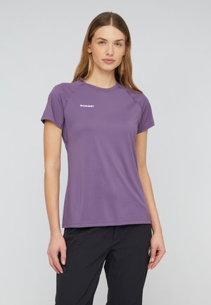 Jeune femme portant un t-shirt violet Mammut et un pantalon noir, debout devant un fond clair uni.