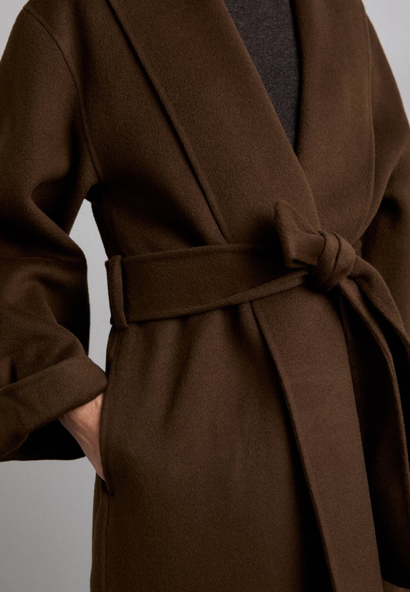 Cappotto di lana marrone con design avvolgente, dotato di una cintura legata, ampi risvolti e polsini arrotolati. La texture appare morbida e calda.