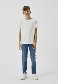 Camiseta de algodón blanca de manga corta combinada con jeans de mezclilla azules. El modelo lleva zapatillas blancas y está de pie contra un fondo liso.