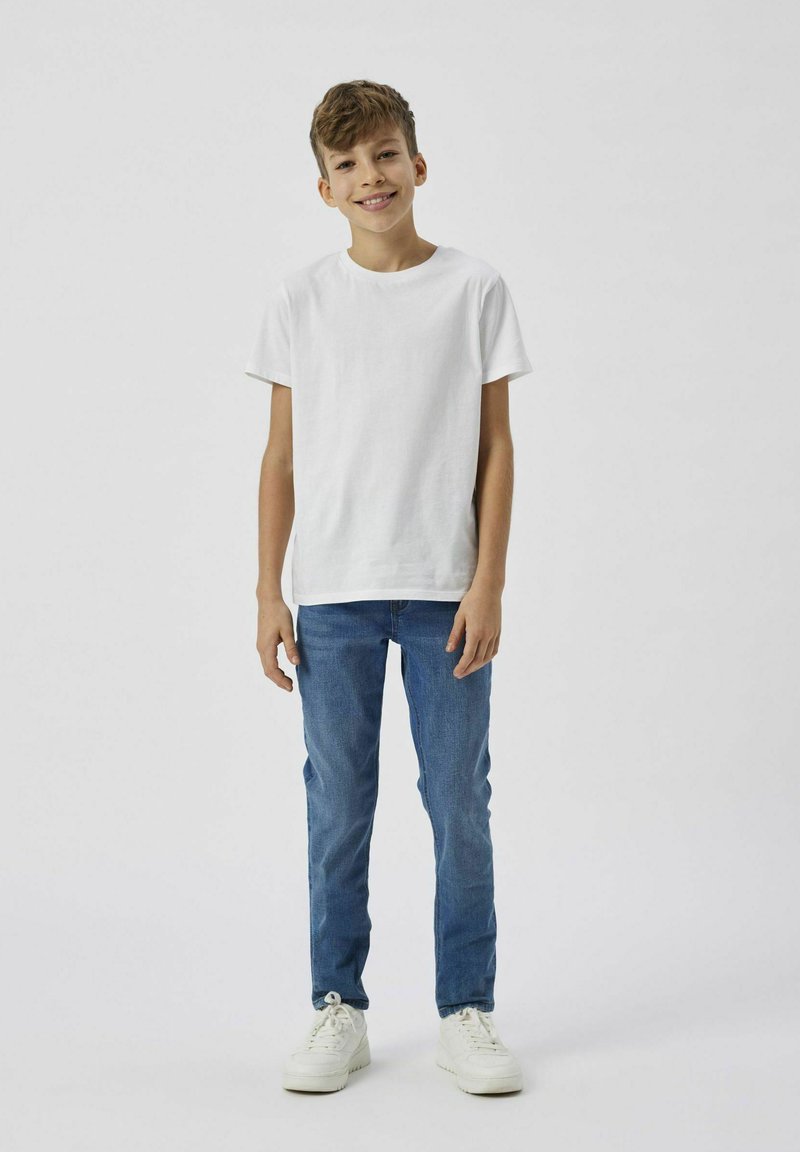 Camiseta de algodón blanca de manga corta combinada con jeans de mezclilla azules. El modelo lleva zapatillas blancas y está de pie contra un fondo liso.