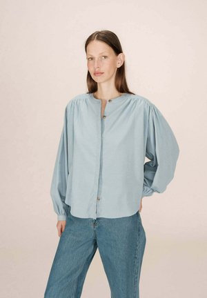 Blouse bleu clair à boutons, avec des manches longues bouffantes et un col rond. Dotée de boutons sur le devant et d'une coupe décontractée. Associée à un jean.