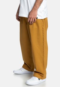 Volné hořčicově žluté kalhoty z bavlněného kepru, s bočními kapsami a rovnými nohavicemi. Nošeny s bílými teniskami.