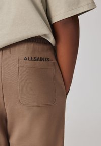 Detail osoby nosící béžové kalhoty s zadní kapsou označenou nápisem "ALLSAINTS" a světle béžové krátké rukávy košile.