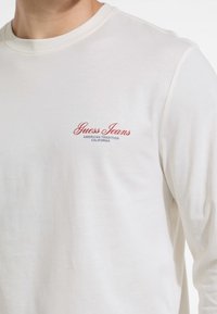 Long-sleeve beige cotton shirt with a crew neck and red "Guess Jeans" logo on the left chest, featuring a smooth, soft texture.