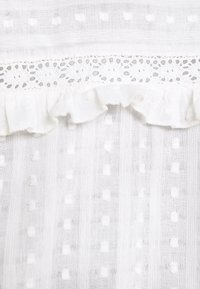 Vitt genomskinligt tyg med små upphöjda prickar, en spetskant och en volangdetalj.