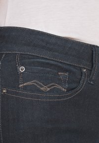 Vaqueros de mezclilla azul oscuro con costuras en los bolsillos, herrajes de cobre y una cinturilla sin costuras, mostrando una vista en primer plano de la textura de la tela.