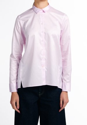 Personne portant une chemise rose clair à manches longues avec un col et un pantalon foncé, debout devant un fond blanc uni.