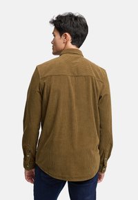 Camisa de pana marrón con rayas verticales, de manga larga y puños abotonados. Presenta un diseño de yugo y un dobladillo redondeado en la parte posterior.