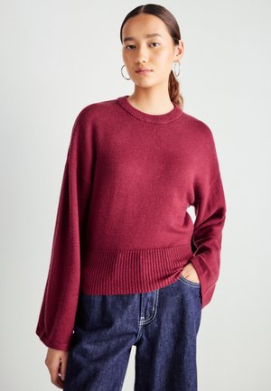 Pull en maille bordeaux avec un ourlet côtelé large et des manches bouffantes, fabriqué en tissu doux. Assorti à un jean bleu foncé à coupe ample.