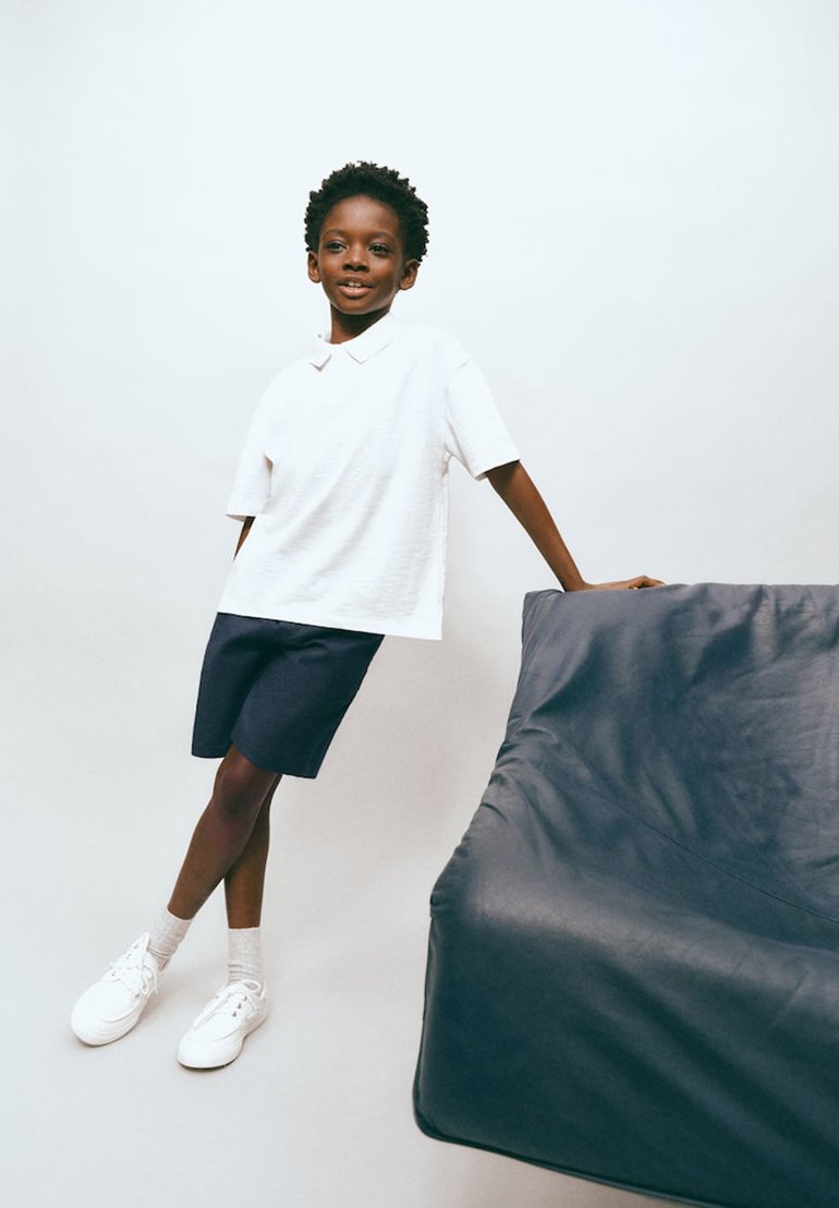 Garçon en polo blanc et short bleu marine appuyé contre un pouf gris foncé sur fond blanc uni.