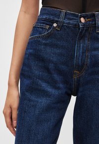 Tumši zili džinsa džinsi ar augstu jostasvietu, izceļoties ar kontrastējošu oranžu šuvumu, priekšējām kabatām un vara pogas aizdari.