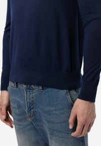 Marineblauer Wollpullover mit einem gerippten Saum und Bündchen, getragen über verblassten blauen Jeans. Der Stoff hat eine glatte Textur und ein schlankes Design.