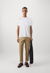 T-shirt en coton blanc avec une poche, chinos beiges, veste foncée à la main, mocassins marron et chaussettes grises, le tout sur un fond uni.
