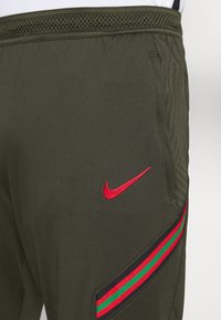 Olivgröna träningsbyxor i slät tyg, med en röd Nike-logotyp, sidofickor och ett randigt mönster med röda och gröna detaljer.