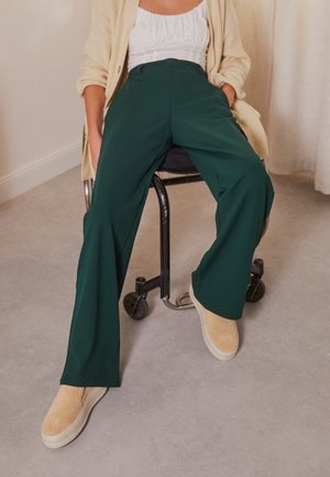Pantalons larges verts en tissu lisse, associés à des chaussures à enfiler beige. La tenue est agrémentée d'un cardigan crème. Assis sur une chaise.