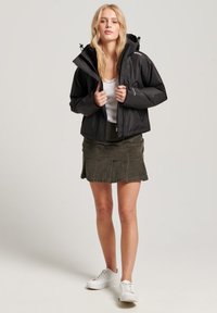 Zwarte waterdichte jas met een rits en een verstelbare capuchon, gelaagd over een witte top. G gedragen met een olijfgroene rok en witte sneakers.