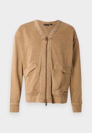 Beežist pehmest kangast lukuga jope, millel on kaks küljetaskut, V-kaelus ning nähtavad õmblusdetailid varrukatel ja alläärmel.