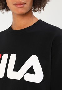 Sudadera negra con cuello redondo acanalado y un gran logo blanco que dice "FILA" con un acento rojo. El material parece suave y texturizado.