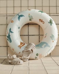 Vihreän ja oranssin pastellisävyisiä merielämän kuvituksia esittelevä kelluva rengas. Beige, teksturoitu sandaali rengasta vasten.