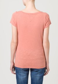 Camisa de color melocotón con mangas cortas, hecha de tela ligera, con un cuello redondo y un detalle en la costura trasera. Jeans visibles por debajo.
