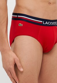 Raudoni medvilniniai apatiniai su mėlynu juosmeniu, kuriame yra baltos ir raudonos juostelės, ir mažas žalias Lacoste logotipas priekyje. Minkštas tekstūra, prigludęs fit.