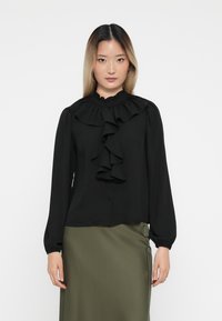Black long-sleeve blouse with ruffled collar and front detailing, featuring button closure and soft fabric. Paired with a green skirt.